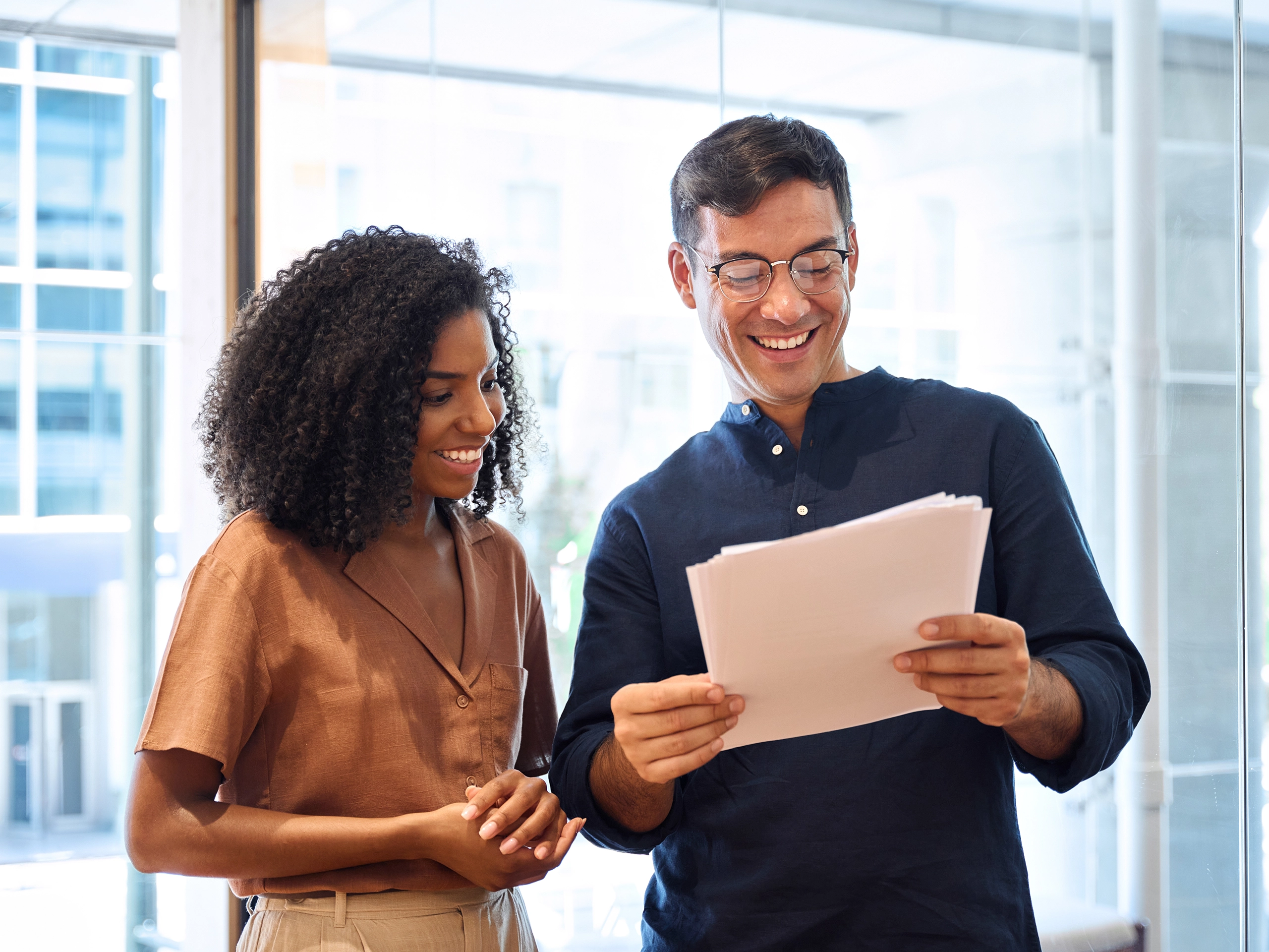 Business team of two happy young busy diverse colleagues, professional business man and business woman executives working together standing in office looking at project documents and talking.