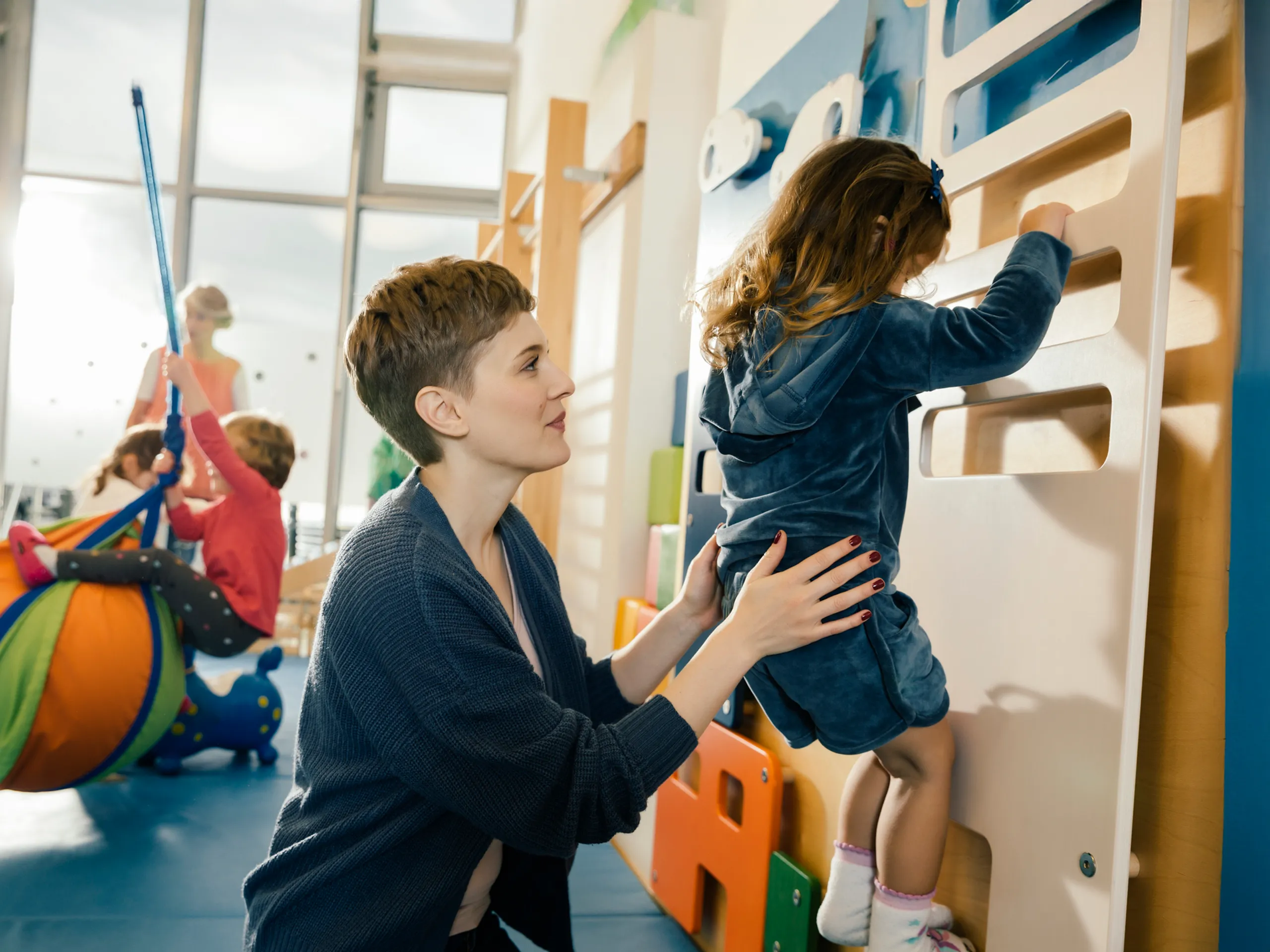 A caregiver helps a child climb a climbing wall in a daycare center.