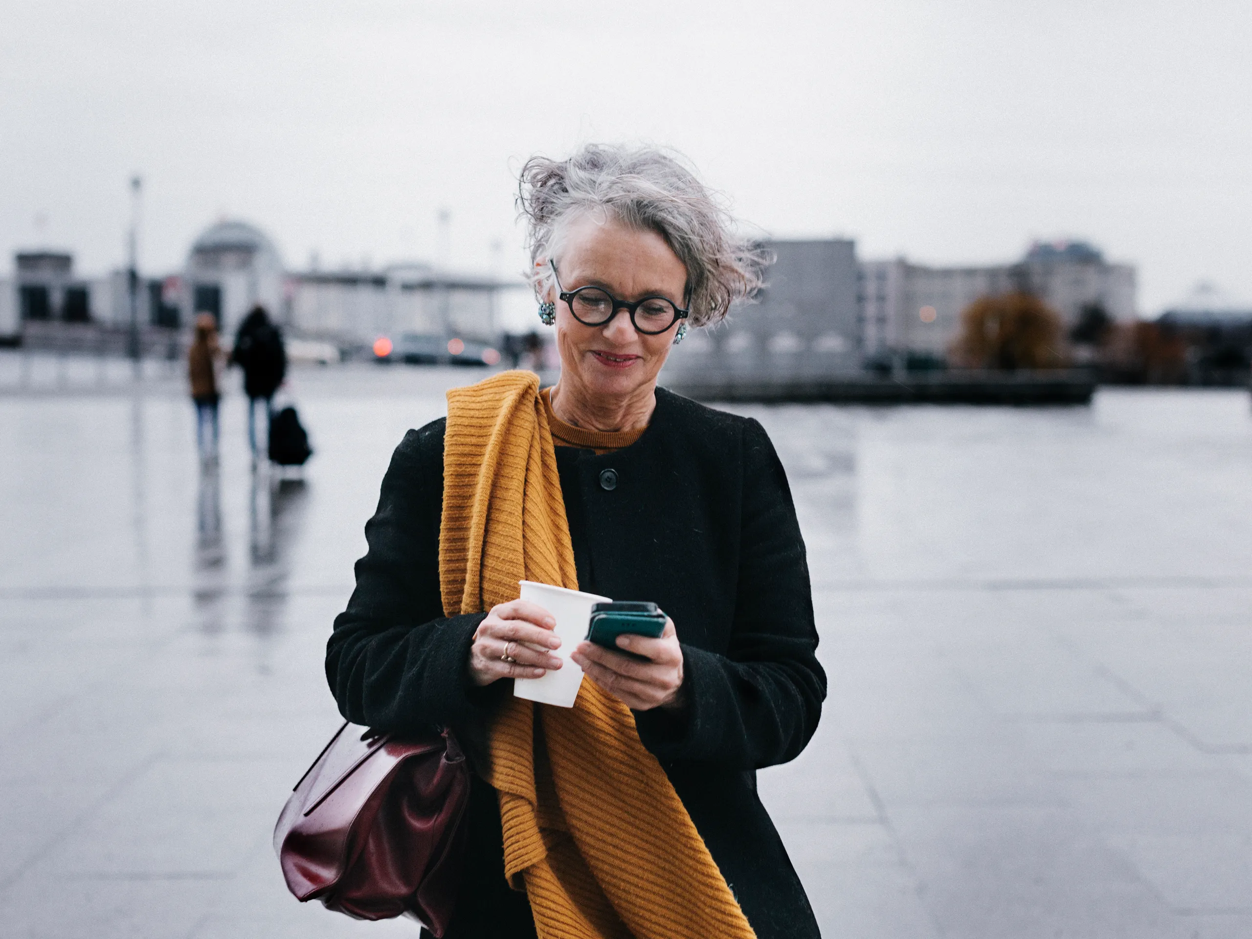 Stylish older woman using smartphone and holding coffee outside.